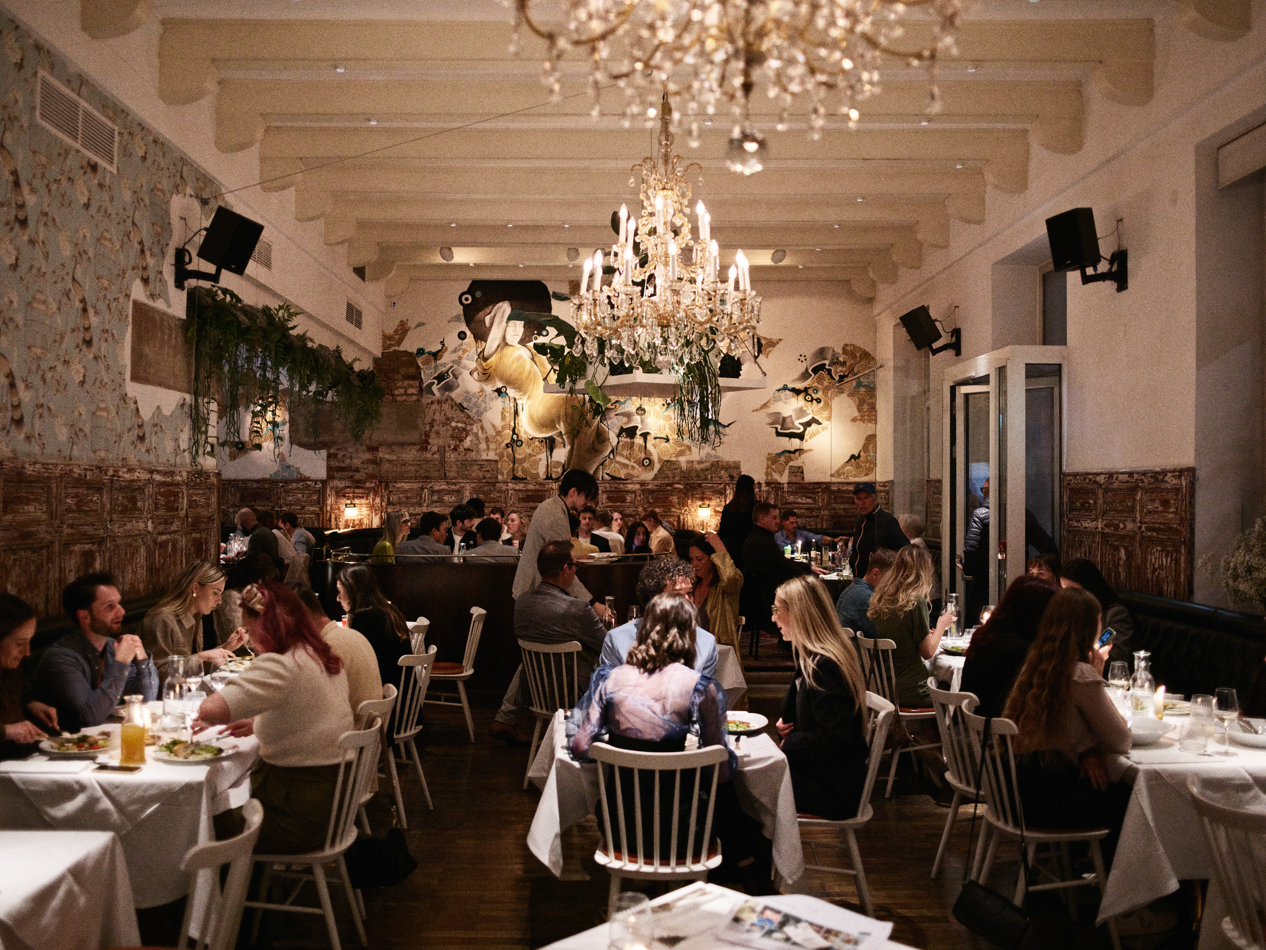 a restaurant filled with people, with chandeliers and plants hanging from the ceiling